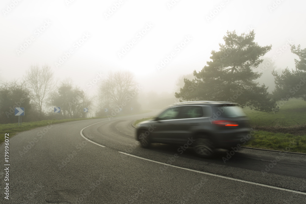Foggy asphalt curved road with cars passing through the forest. Weather with low visibility in the region of Normandy, France. Country landscape on misty day. Toned