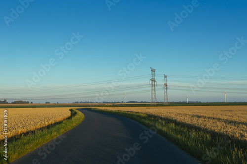 High voltage power lines and transmission towers. Asphalt road passing through agricultural fields in Normandy, France. Electric power industry, road network and agriculture concept