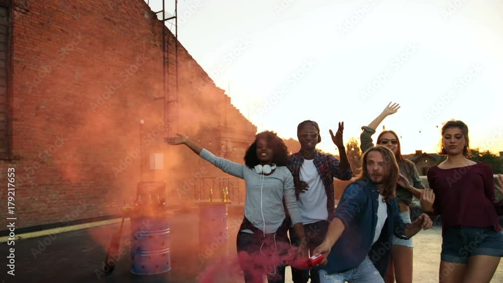 Young attractive people walking and playing with pink smoke grenade at the rooftop party in urban space. The sunset background. Outside. Multiethnical Young attractive people walking and playing with pink smoke grenade at the rooftop party in urban space. The sunset background. Outside. Multiethnical