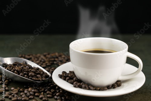 Hot coffee in the white cup with roast coffee beans and scoop on stone table in black background