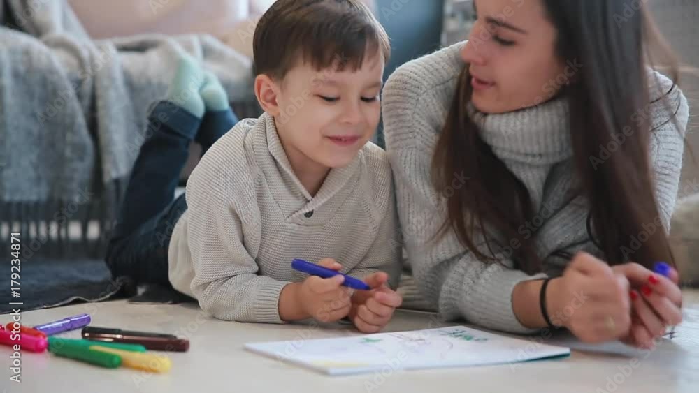 Beautiful young mother in a warm sweater lying on the floor with my son drawing with markers on paper portraying his family. The child learns to draw. Close-up Beautiful young mother in a warm sweater lying on the floor with my son drawing with markers on paper portraying his family. The child learns to draw. Close-up