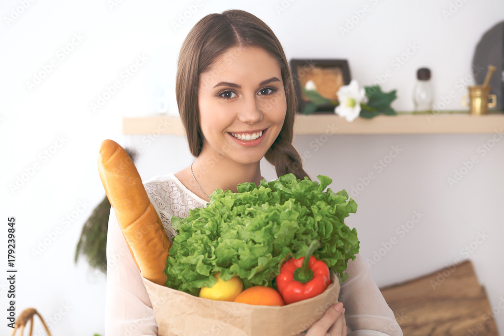 Young happy woman holding paper bag full of vegetables and fruits while smiling. Girl have made shopping and ready for cooking in kitchen Young happy woman holding paper bag full of vegetables and fruits while smiling. Girl have made shopping and ready for cooking in kitchen