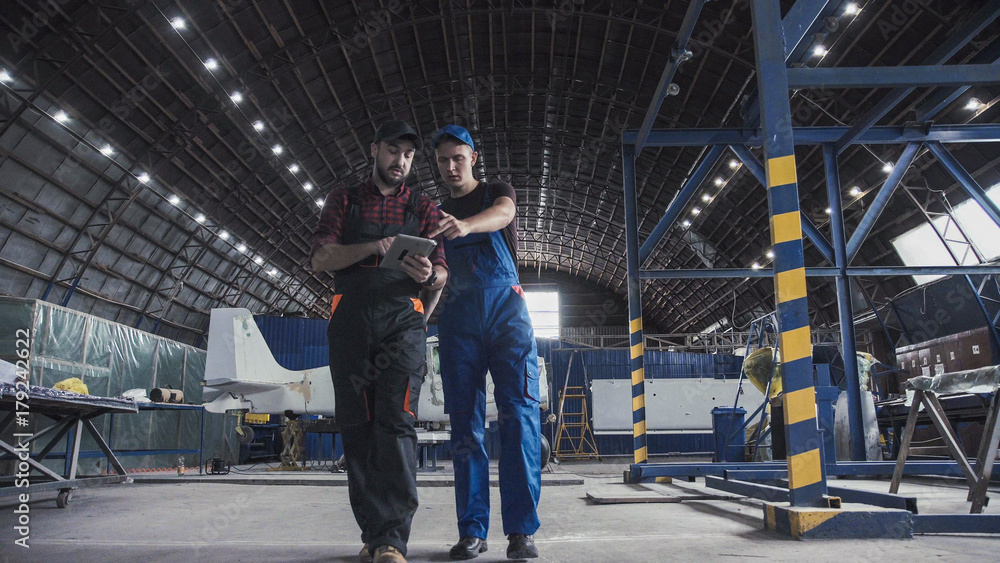 Two flight engineers walking through a large aircraft hangar talking and gesturing together - obrazy, fototapety, plakaty 