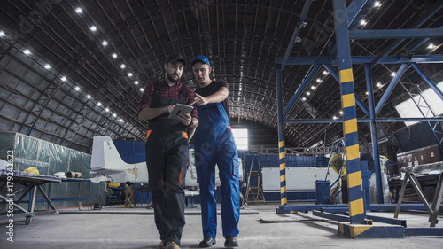 Two flight engineers walking through a large aircraft hangar talking and gesturing together