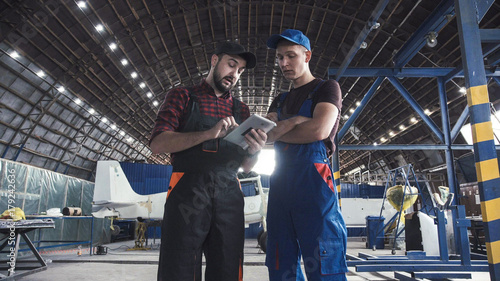 Two flight engineers walking through a large aircraft hangar talking and gesturing together
