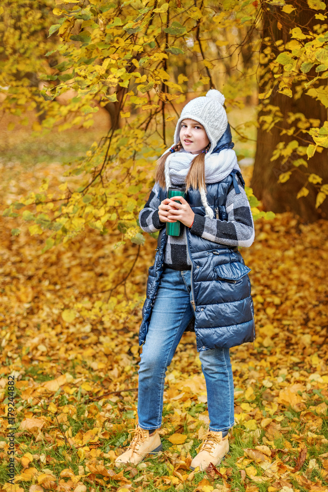 cute teenager girl in a white hat and volumetric scarf holding a thermos with tea in an autumn park