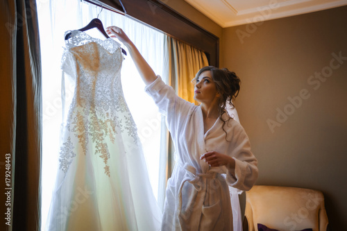 Bride hanging wedding dress on a hanger near window