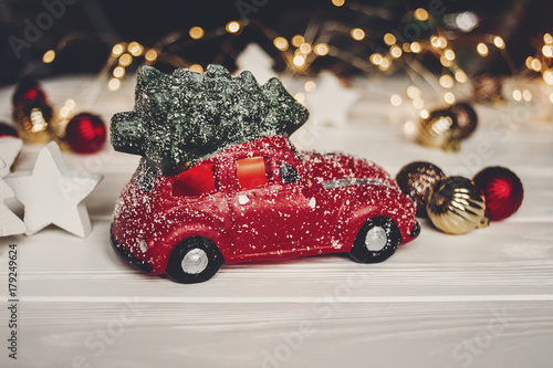 red car toy with christmas tree on top and simple ornaments on white wood with lights in background, space for text. christmas present. seasonal greetings. happy holidays. xmas gift
