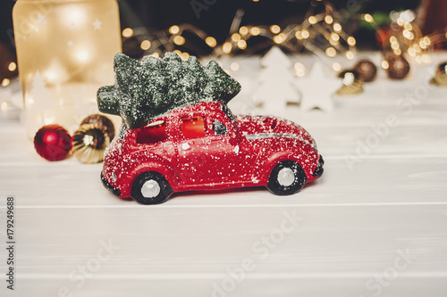 christmas present. red car toy with christmas tree on top on white wood with lights in background, space for text. seasonal greetings. happy holidays. xmas gift. holiday celebration