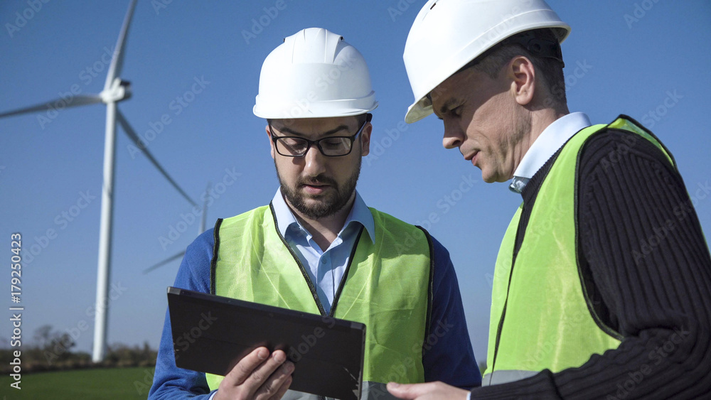 Two engineers wearing hard hat discussing over digital table against turbines on wind farm Two engineers wearing hard hat discussing over digital table against turbines on wind farm