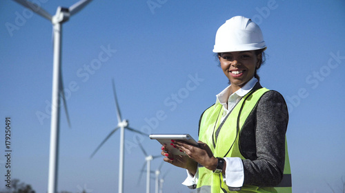 Female engineer wearing hard hat standing with digital tablet against wind turbine on sunny day and looking at camera smilingly