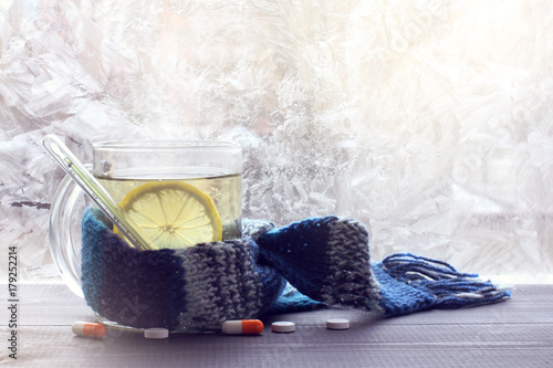treatment of colds in winter/ hot infusion with lemon in mug, wrapped in a scarf with a thermometer on the background of pills and frozen windows