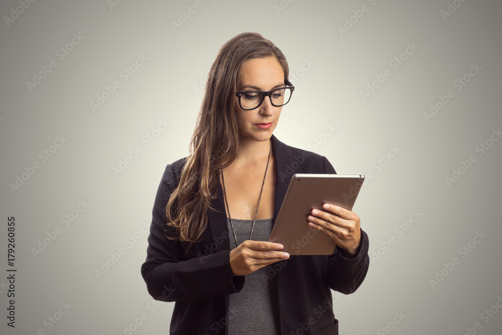Imagem de mulher linda intelectuel de oculos em pé enquanto segura o comprimido digital em suas mãos e lendo o texto ao fundo cinza. Chefe Mulher de negócios que trabalha no escritório. Imagem de mulher linda intelectuel de oculos em pé enquanto segura o comprimido digital em suas mãos e lendo o texto ao fundo cinza. Chefe Mulher de negócios que trabalha no escritório.