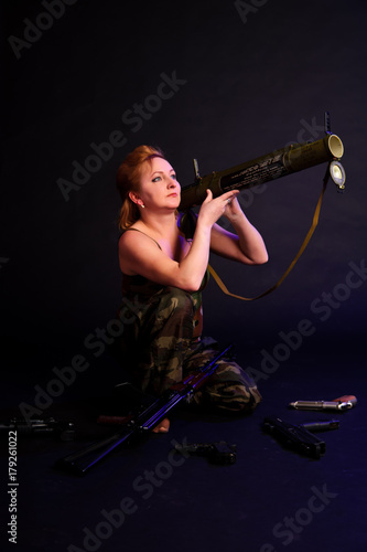 A middle aged, white, woman holds a RPG18 Mukha grenade launcher, with weapon around against black photo