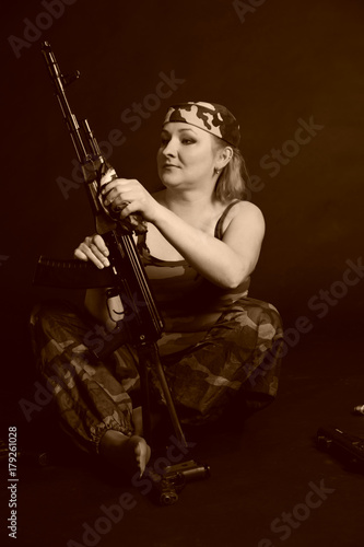 A middle aged, white, woman holds an AK47 automatic rifle, with weapon around against black photo