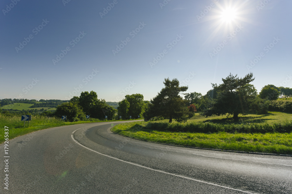 Empty asphalt curvy road passing through green fields and forests. Countryside landscape on a sunny spring day in France. Sunbeams in the sky. Transport, industrial agriculture, road network concept