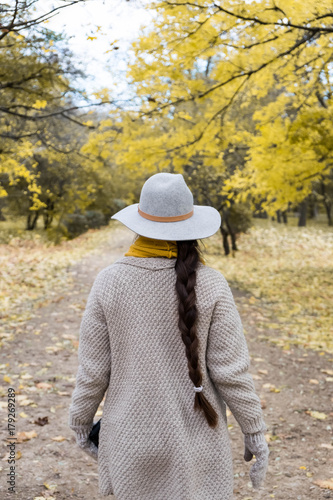 Rear view of brunette girl in autumn fall park in grey hat, woolen handmade cardigan. Back view of autumn portrait of woman outdoors