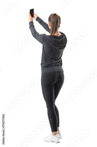 Rear view of sporty athletic woman taking selfie while holding cellphone in both hands. Full body length portrait isolated on white background.