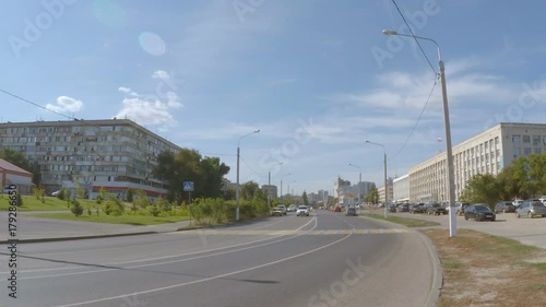 Cars are driving down the street of the city between the buildings. Sunlight and blue sky with white clouds.