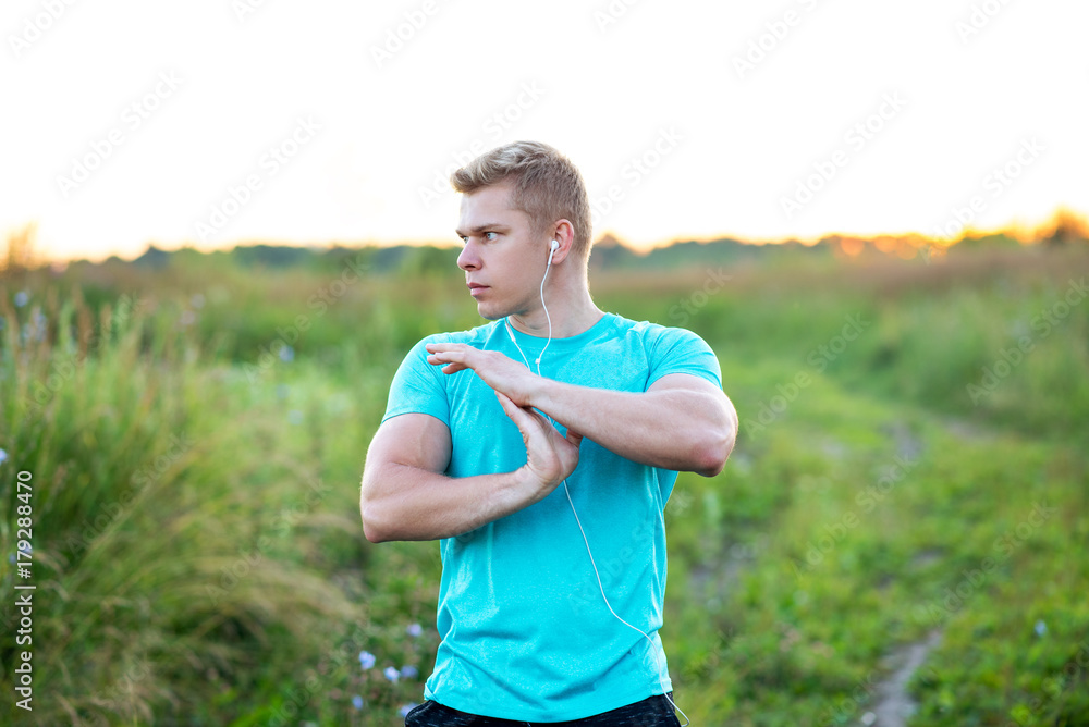Warm-up hands, a young guy runner early in the morning doing the headphones. A man in the park listens to music, a confident look. In summer, life style, motivation is warming up. Outdoors in the city Warm-up hands, a young guy runner early in the morning doing the headphones. A man in the park listens to music, a confident look. In summer, life style, motivation is warming up. Outdoors in the city