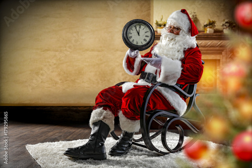 Santa Claus sitting in a wooden rocking chair. In the foreground is a fuzzy Christmas tree with baubles and lights. On the third floor is a wall with a fireplace. 