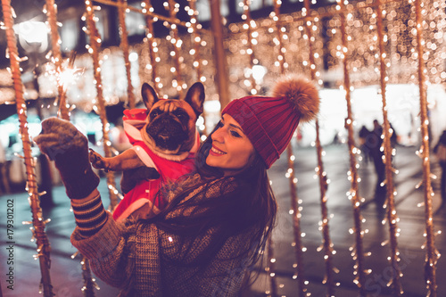 Beautiful young brunette woman with French bulldog enjoying Christmas or New Year night on a city street. 