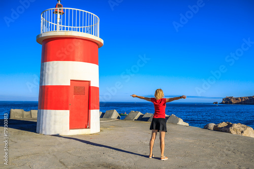 Happy blonde woman with open arms at red lighthouse of Porto de Abrigo from Nazare in Central Portugal, Europe. Female tourist admires the high cliffs of Nazare Sitio and its fortress on background.