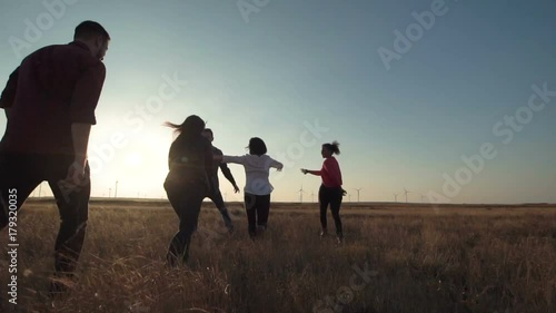 Group of friends running through field towards wind farm at sunset