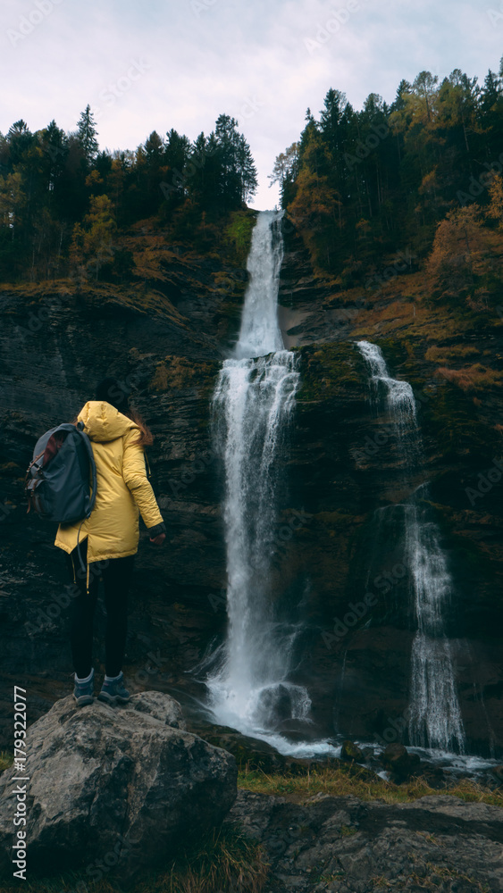 Young Caucasian female hiker in yellow raincoat wearing backpack enjoys the view of a beautiful waterfall in French Alps