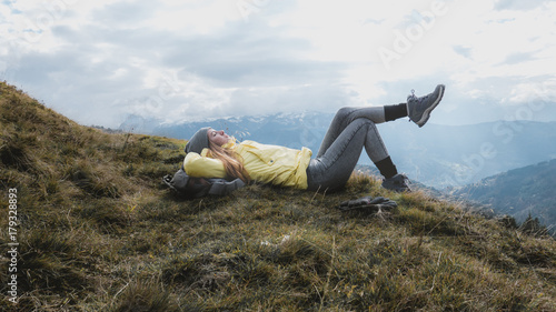Hipster young girl with backpack hiking in mountains. Happy tourist traveler lying on the ground and enjoying the view from the top of the mountain