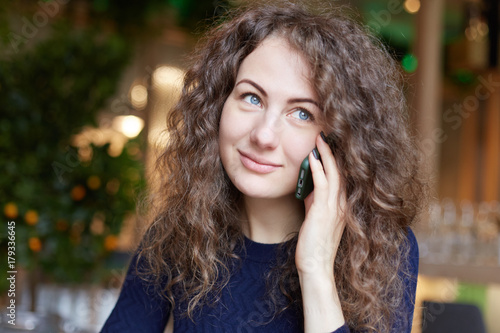 Pretty blue eyed woman with dreamy look holding smartphone communicating with shining smile, sitting in modern cafe, being happy to hear her friend. People, modern technologies, communication concept.