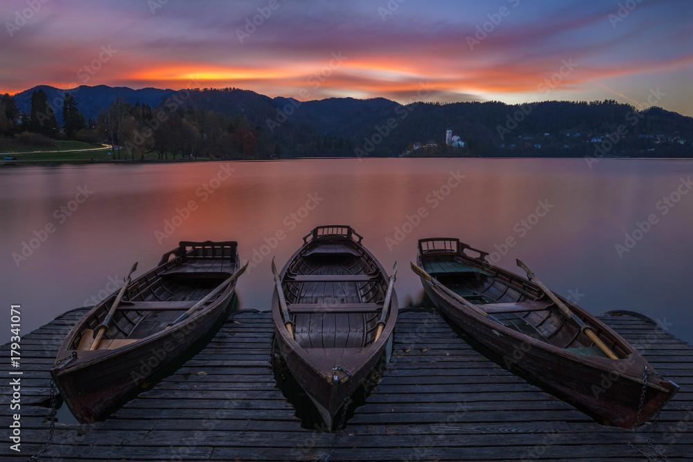 Bled, Slovenia - Traditional boats at Lake Bled with beautiful dramatic sunset and the Assumption of Mary Pilgrimage Church and mountains at background at sunset