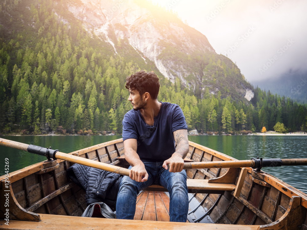 Young casual man sitting in boat and rowing while looking away on background of lake and mountains.
