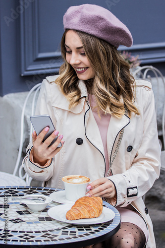 Receiving good news. Vertical shot of beautiful blonde woman in beret looking at phone and smiling while sitting in french vintage cafe. Woman drinking coffee with croissant and using smart phone.