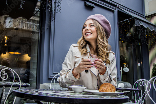Cheerful french woman. Low-angle view of beautiful blonde woman in beret looking away and smiling while sitting in french vintage cafe. Woman drinking coffee with croissant. French breakfast concept. photo