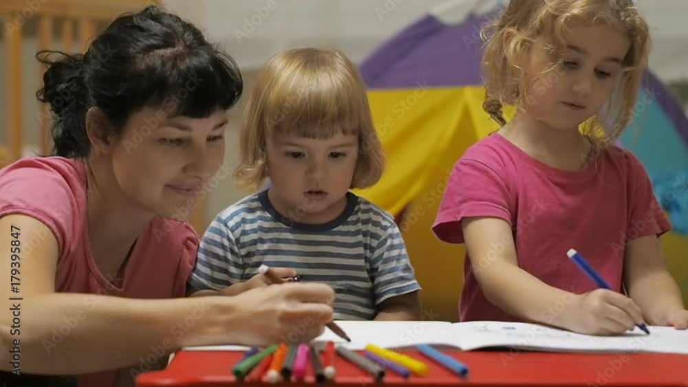 The educator and two little children girl and boy sitting at table drawing. Brother and sister and their mother drawing with colorful crayons and markers The educator and two little children girl and boy sitting at table drawing. Brother and sister and their mother drawing with colorful crayons and markers
