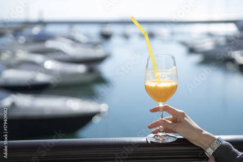 Female hand holding a glass of orange juice on the background of boats