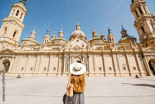 Young woman tourist in sunhat standing back in front of the famous cathedral on the central square during the sunny weather in Zaragoza city, Spain