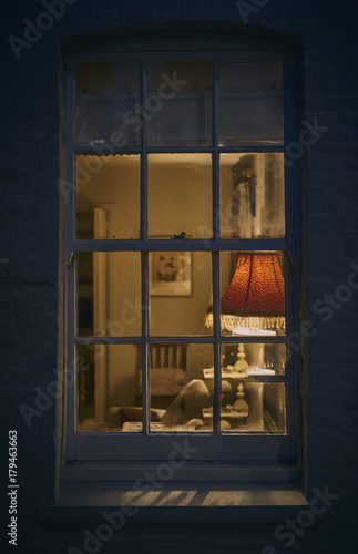 Looking through a window at night from outside into a rustic cottage with lamp
