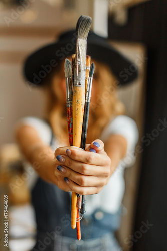 Beautiful long hair artist girl, in a black hat and denim dangarees holding brushes before the easel photo