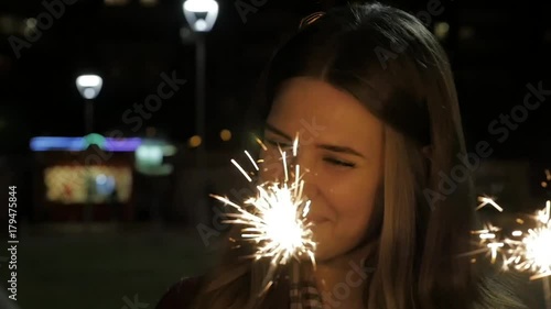 Smiling teenage girl on the street at night with sparklers. Young woman celebrating an event the New Year is coming. Portrait of girl holding sparkles in slow motion photo