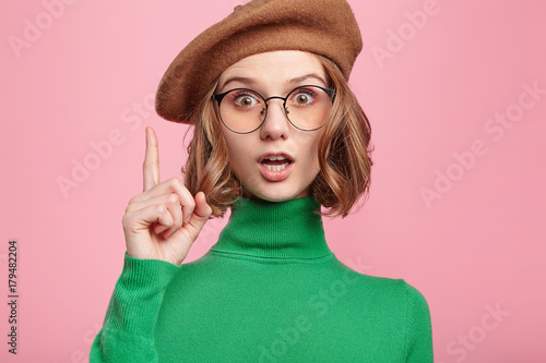 Adorable beautiful young woman gets good idea in mind how improve project, raises fore finger, wants to sound and express thoughts, has worried expression, doesn`t want to forget brilliant thought