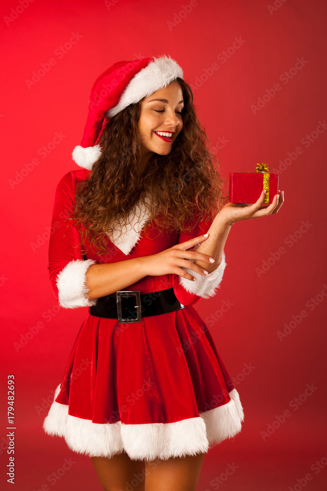 Beautiful young woman in Santa dress holds a present over red background Beautiful young woman in Santa dress holds a present over red background