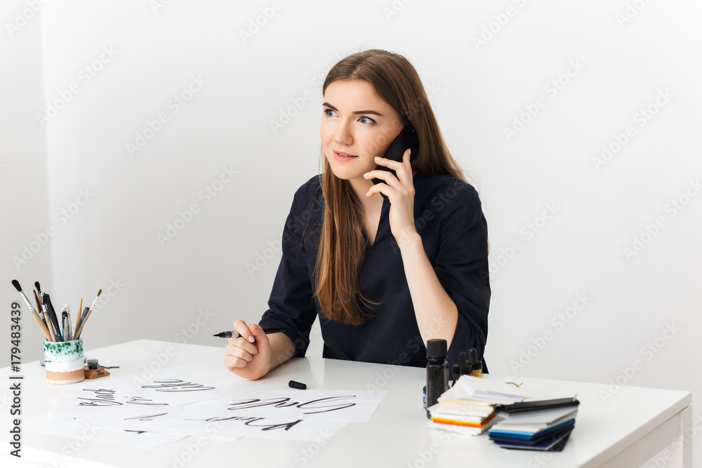 Beautiful photo of young lady sitting at the white desk and talking on her cellphone while happily looking aside isolated Beautiful photo of young lady sitting at the white desk and talking on her cellphone while happily looking aside isolated