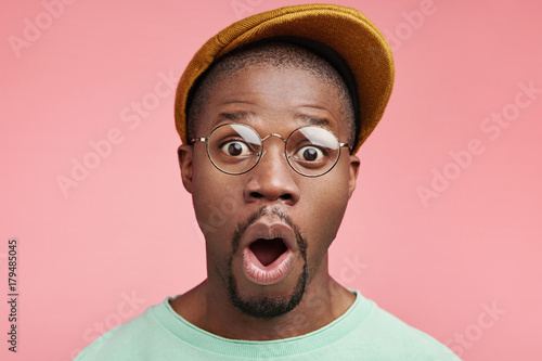 Fashionable black male with specific appearance keeps mouth widely opened, stares at camera, being shocked with prices in shopping mall, wants to buy new suit. Emotional drk skinned man indoor © wayhome.studio 