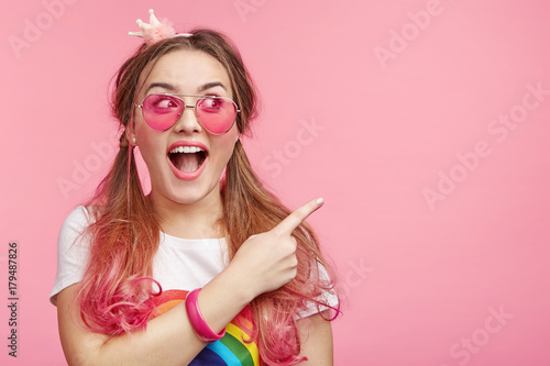 Cute astonished young female with pony tails and pink hair tips, points at copy space with fore finger advertises something, keeps mouth widely opened. People, advertising, surprisment concept photo