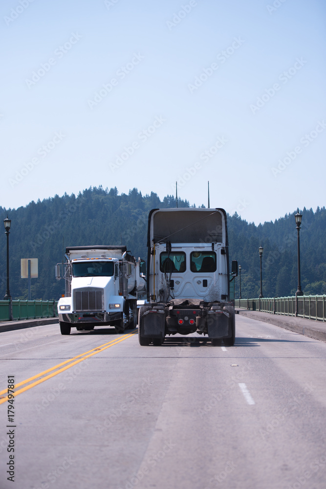 Two big rigs semi trucks dumper and semi tractor are moving towards each other on the way to the bridge