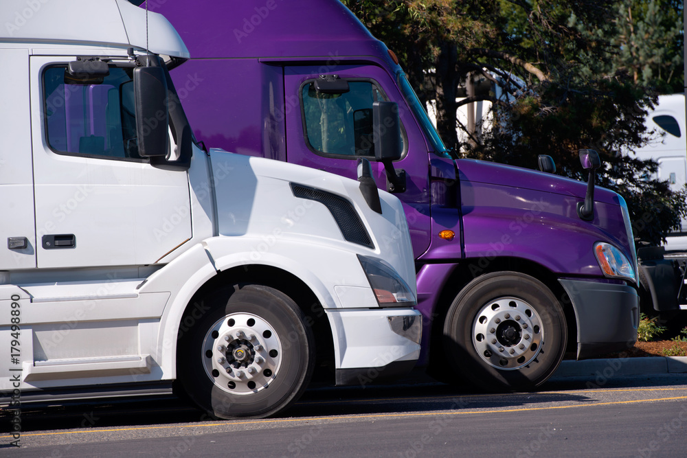 White and purple big rigs semi trucks stand on truck stop side by side