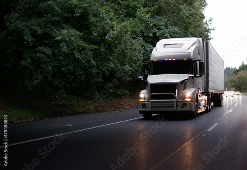 Modern big rig semi truck with guard and turn on headlights and reefer semi trailer on evening road photo