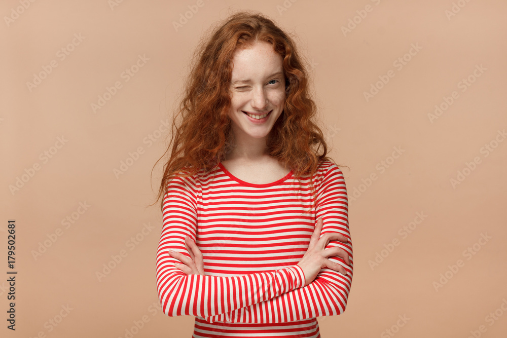 Pretty curly redhead girl in striped t-shirt with crossed arms smiling and winking, looking at camera Pretty curly redhead girl in striped t-shirt with crossed arms smiling and winking, looking at camera